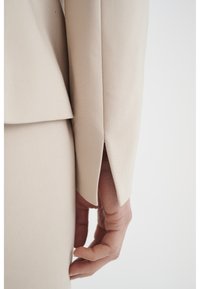 Beige upravená sako rukáv s ventilačními detaily, ukazující hladkou texturu a čisté linie. Ruka umístěná poblíž oděvu.