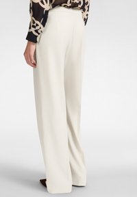Pantalones de pierna ancha en color crema. Presentan una cinturilla ajustada y una tela fluida. Textura suave y detalles de diseño mínimos. Calzado parcialmente visible.