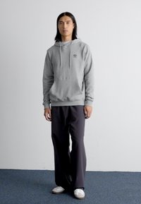 Felpa grigia con tasca frontale e cordini, abbinata a pantaloni neri larghi e sneakers bianche, in piedi su un pavimento blu con moquette.