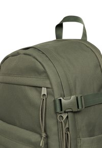 Mochila de tela verde con un acabado texturizado, que cuenta con un bolsillo frontal, tiradores de cremallera y un asa de cinta. Acentos oscuros complementan el diseño.
