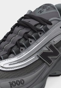 Γκρι αθλητικό παπούτσι New Balance 1000 με μαύρα κορδόνια, ασημί ρίγα και υφασμάτινη υφή στη λεπτομέρεια στο πλάι και τη γλώσσα.