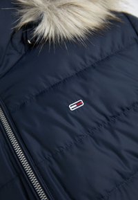 Navy blue puffer jacket featuring a fur-lined hood, silver zipper, and a small logo with red and white accents on the front.