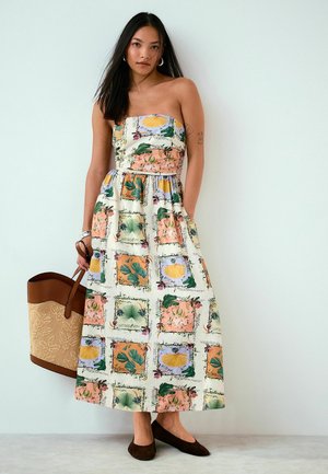 Frau trägt schulterfreies Maxikleid mit Blumen-Patchwork, hält große beige-braune Tragetasche und steht vor weißer Wand.