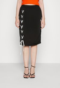 Donna che indossa una gonna nera a lunghezza ginocchio con dettagli in pizzo bianco sui lati, un top arancione e sandali neri con tacchi alti e cinturini.