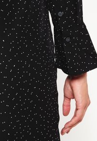 Černý oděv s bílými puntíky, který má manžetu na knoflíky. Tkanina vypadá hladce s mírným leskem a dominuje jí elegantní střih.