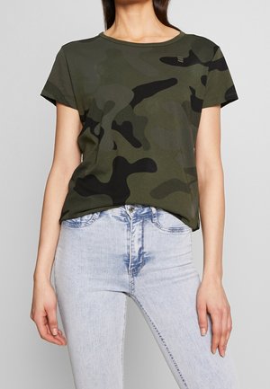 Femme portant un t-shirt manches courtes camouflage vert et noir et un jean taille haute bleu clair.