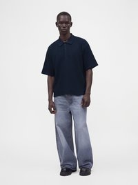 Hombre que lleva una camiseta polo azul marino de manga corta, vaqueros anchos de pierna en azul claro y zapatos negros gruesos, de pie sobre un fondo claro.
