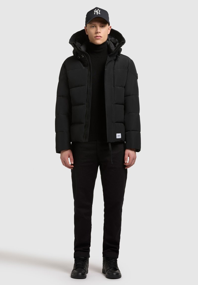 Chaqueta de plumas negra con cremallera delantera y capucha, llevada sobre un suéter de cuello alto negro y pantalones negros. Acentuada con una gorra negra. Textura suave.