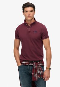 Bordeaux polo skjorte med knapkrave og broderet logo, parret med blå jeans og en bundet ternet flannel skjorte omkring livet.