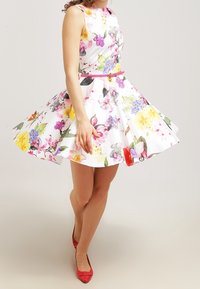 Robe fleurie à fond blanc, ornée de fleurs colorées. Un corsage ajusté mène à une jupe évasée. Accentuée d'une ceinture rose et portée avec des chaussures rouges.