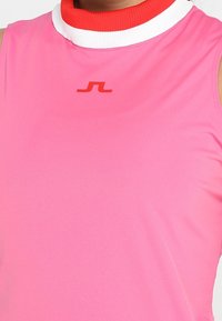 Bezrukávový sportovní top v jasně růžové látce, s vysokým červeným a bílým žebrovaným límcem a malým logem na hrudi.