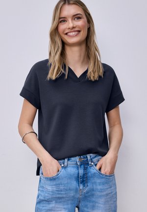 Mujer sonriente con cabello rubio, vestida con una camiseta oscura de manga corta y cuello en V, y jeans azul claro, con las manos en los bolsillos.