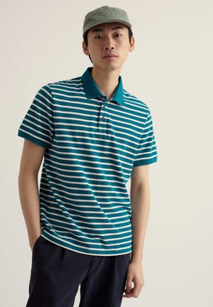 Uomo con polo a righe verde e bianca, pantaloni scuri e cappellino verde chiaro, in piedi davanti a uno sfondo semplice color avorio.