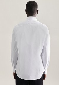 Chemise blanche à manches longues avec un col classique, réalisée en tissu lisse. Présente un ourlet arrondi et des poignets à boutons, avec des détails de couture minimaux.