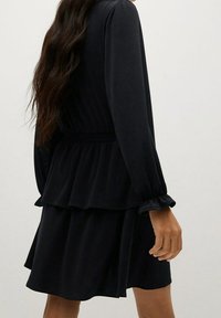 Vestido negro con mangas largas y abullonadas, borde con volantes en capas y cintura fruncida, con una suave tela texturizada.