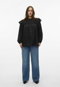 Blusa nera a volant con pois testurizzati, caratterizzata da un collo alto e maniche lunghe, abbinata a jeans in denim blu a gamba larga e tacchi neri.