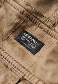 Beige stof med en tekstureret overflade, prydet med et sort mærke med hvid tekst, der læser "SUPERDRY Manufacturing Co." indrammet i en rektangulær form.