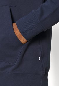 Mörkhyad hand insatt i framfickan på en marinblå sweatshirt med ribbad mudd och liten vit tygetikett.