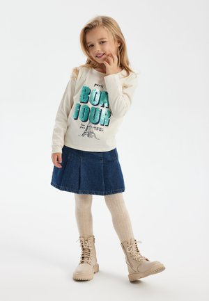 Lichtcrème langemenig shirt met een "BONJOUR" grafiek in teal sequins, gecombineerd met een donkere denim rok en beige veterschoenen.