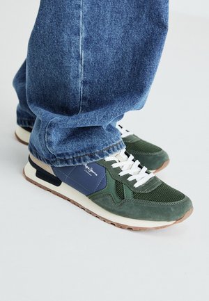 Sneakers verdi e blu navy con dettagli in rete e suede, lacci bianchi e suola beige. Indossati con jeans in denim blu, mostrando la zona della caviglia.