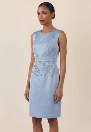 Frau trägt ein ärmelloses hellblaues Kleid mit 3D-Blumenapplikationen vorne und einem knielangen Saum vor einem beigen Hintergrund.