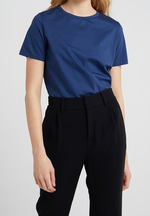 Femme portant un t-shirt bleu uni à manches courtes, rentré dans un pantalon noir taille haute, sur un fond neutre.