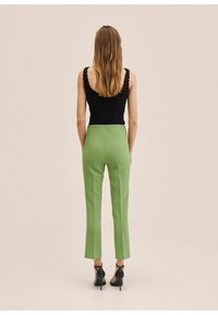 Vrouw met lang haar, gekleed in een zwarte mouwloze top en felgroene op maat gemaakte broek, staande tegen een effen beige achtergrond.