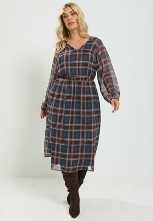 Vrouw met blond haar draagt een lange mouw bruine en blauwe geruite midi-jurk met elastische taille en donkere kniehoge laarzen.
