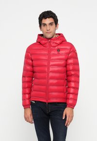 Young man wearing a red quilted hooded jacket and dark jeans, standing against a plain white background.