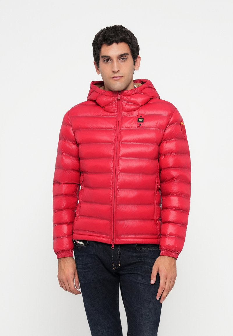 Young man wearing a red quilted hooded jacket and dark jeans, standing against a plain white background.