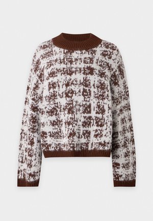 Personne debout portant un pull ample marron et blanc à motifs, un pantalon large foncé et des chaussures marron sur un fond clair uni.