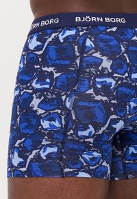 Blue boxer briefs featuring a graphic pattern of overlapping shapes in varying shades of blue and gray, with a solid blue waistband.