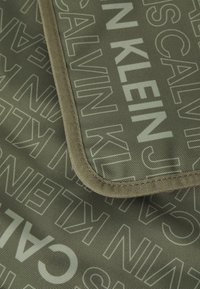 Zelená textilná taška s opakujúcim sa vzorom loga "Calvin Klein" v svetlo sivej farbe, so svetlohnedým lemovaním okrajov. Viditeľná odolná textura.