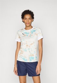Kortermet idrettsskjorte i lys kremfarge med turkis og beige abstrakte mønstre, kombinert med marineblå shorts; har rund hals og logo.