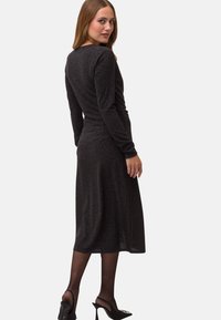 Schwarzes, knielanges Kleid mit langen Ärmeln und tailliertem Schnitt. Weicher, strukturierter Stoff mit dezentem Glanz. Tragen mit hochhackigen Schuhen.