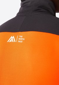 Black and orange jacket with a textured surface, featuring a logo in white on the back, high collar, and color-blocked design.