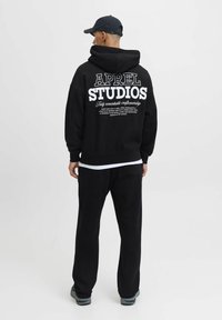 Sort hættetrøje med hvid tekst "APREL STUDIOS" på ryggen, parret med matchende sorte bukser og en sort kasket. Blødt stof, afslappet pasform.