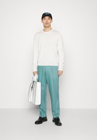 Weißes Langarmhemd, türkisfarbene Hose, weiße Tasche mit schwarzen Akzenten. Schwarze Mütze mit blauer Grafik, schwarze Schuhe. Einfarbiger Hintergrund.