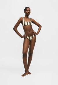 Femme debout, mains sur les hanches, portant un bikini rayé verticalement vert et blanc sur un fond gris clair uni.