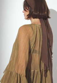 Vestido olivo plisado y transparente con mangas fruncidas, acompañado de una bufanda marrón atada en la parte de atrás. La tela texturizada realza la profundidad visual.