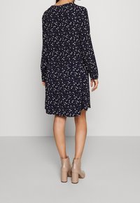 Mörkblå klänning med vitt blommönster, långa ärmar och avslappnad passform. Midilängd, tillverkad av lättviktsmaterial, kombinerad med beige klackstövlar.