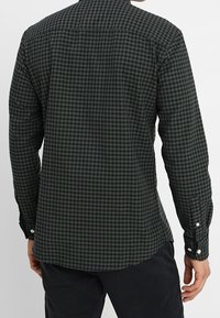 Muž oblečený v tmavě zelené a černé kostkované košili s dlouhým rukávem a černých kalhotách, zobrazený zezadu.