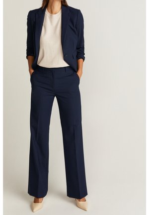 Kvinde iført navyblå blazer og matchende viddebukser med beige top og spidse beige hæle, stående med hænderne i lommerne.