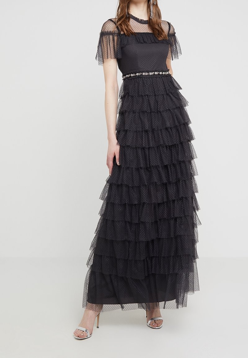 Vestido maxi negro con capas de malla con lunares, que presenta un cuerpo ajustado, mangas con volantines y una cintura adornada con joyas. Tacones de tiras plateados.
