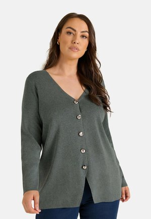 Woman with long brown hair wearing a dark green buttoned cardigan and dark blue pants against a plain light background.