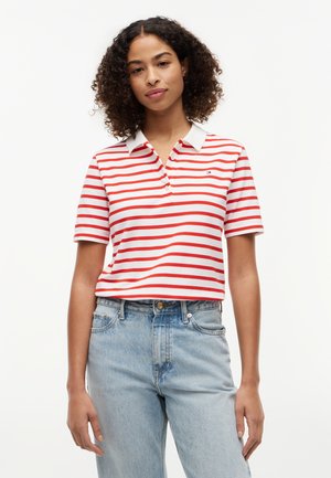 Kvinde med krøllet hår iført en hvid og rødstribet kortærmet polo-shirt og lyseblå denimjeans, stående mod en ensfarvet baggrund.