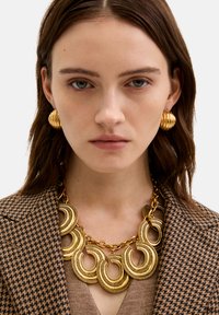 Kullanılmış desenli kahverengi blazer ile birlikte takılan, dairesel halkalar ve karmaşık detaylarla süslenmiş altın tonlamalı kolye, ribbed altın küpelerle eşleştirilmiştir.