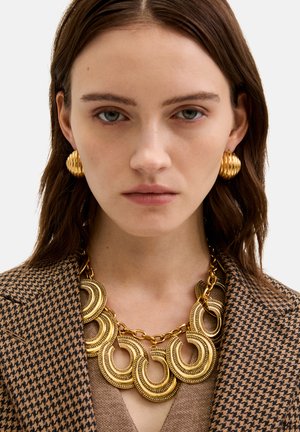 Złota naszyjnik z okrągłymi ogniwami i misternymi detalami, zestawiony z prążkowanymi złotymi kolczykami, noszony z wzorzystym brązowym żakietem.