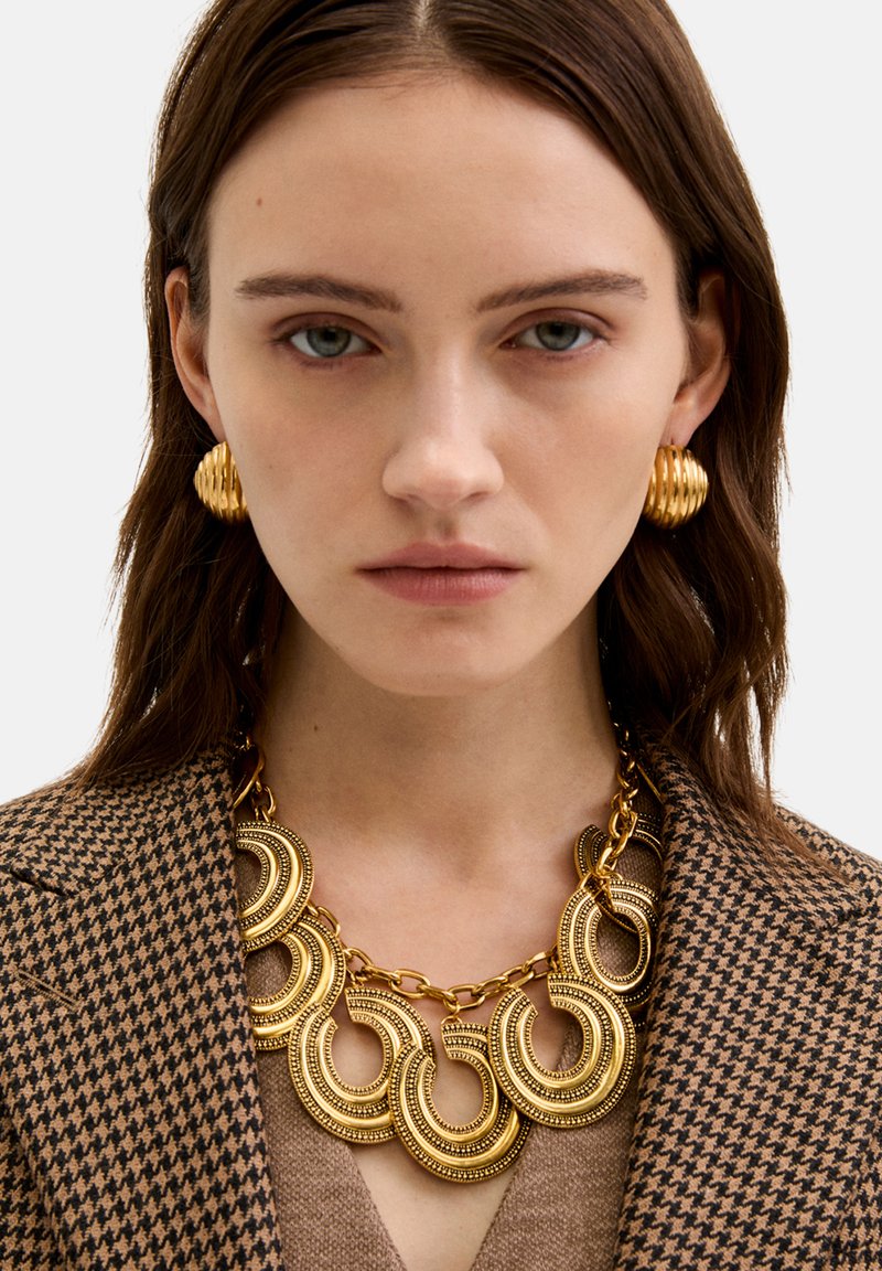 Kullanılmış desenli kahverengi blazer ile birlikte takılan, dairesel halkalar ve karmaşık detaylarla süslenmiş altın tonlamalı kolye, ribbed altın küpelerle eşleştirilmiştir.