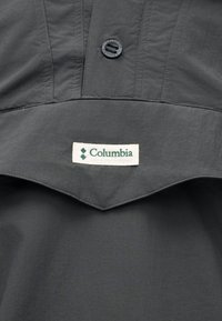 Gros plan sur un tissu gris avec un bouton et une étiquette blanche indiquant Columbia Sportswear Company sur un vêtement d'extérieur.